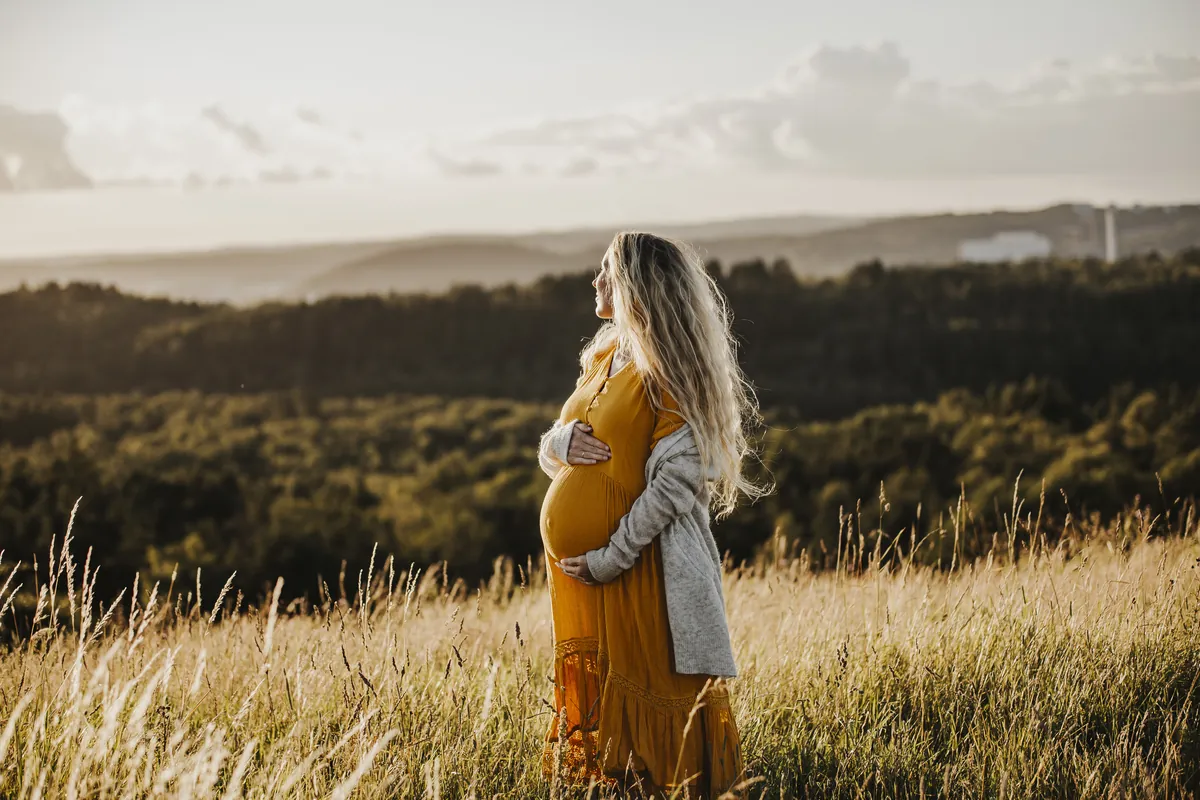 Gravide Mia i oransje kjole og jakke ser utover landskapet i solnedgang