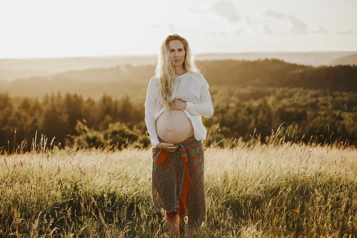 Gravide Mia i hvit skjorte og mønstret skjørt på en eng med utsikt