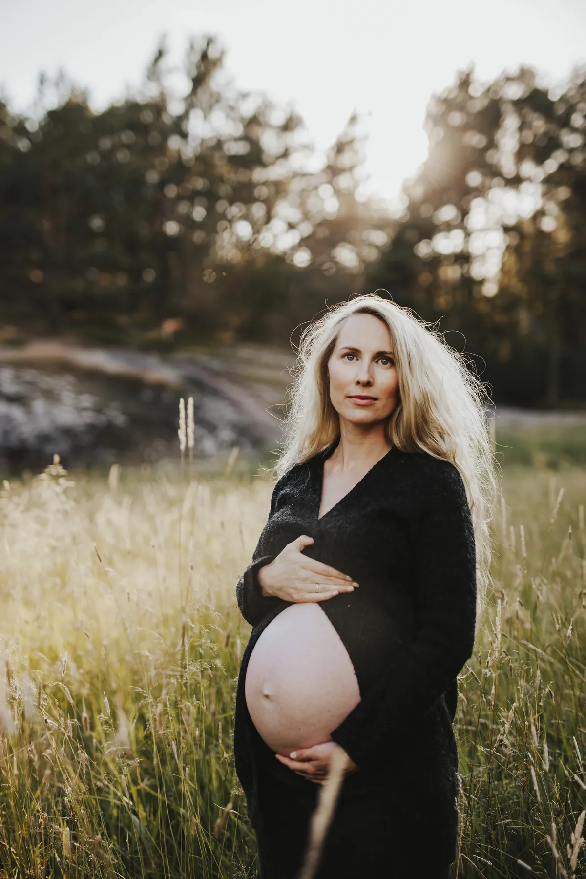 Gravide Mia i svart kjole står rolig i gresset foran skogen