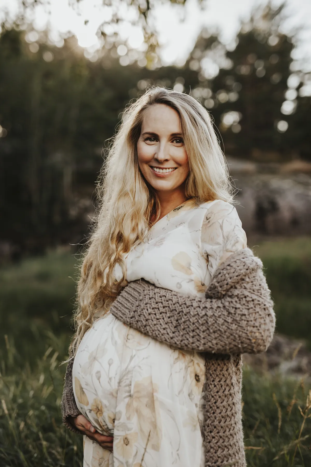 Mia smiler i blomstrete kjole og strikkejakke, gravid, i skogen
