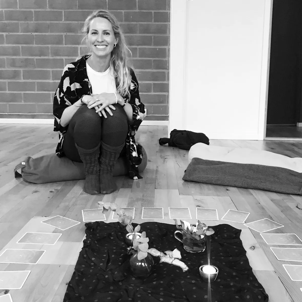 Mia Ruud sitter smilende på gulvet foran en yogasirkel med lys, blomster og kort