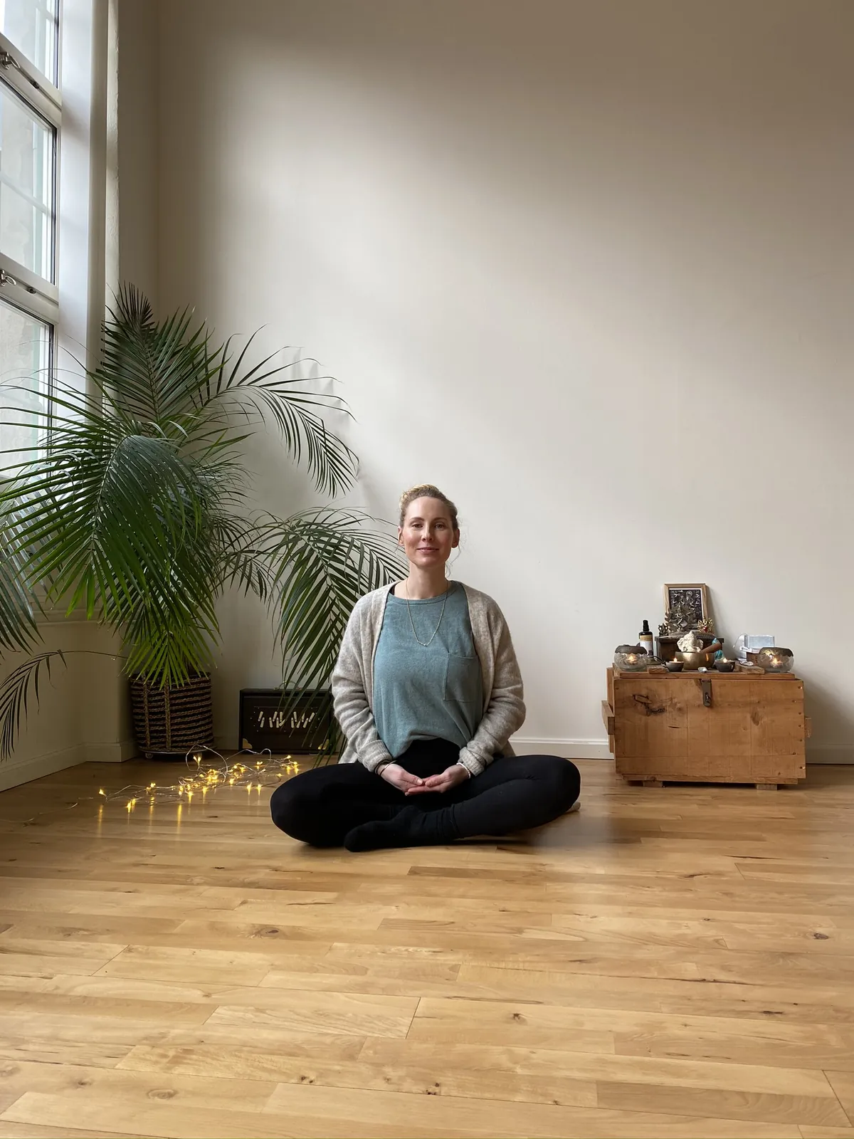 Mia sitter med beina i kryss på gulvet i et lyst yogastudio med palme og lysslynge