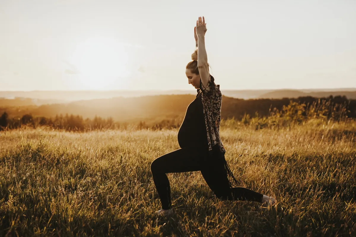 Gravide Mia gjør yoga i lav utfallsstilling på en eng i solnedgang